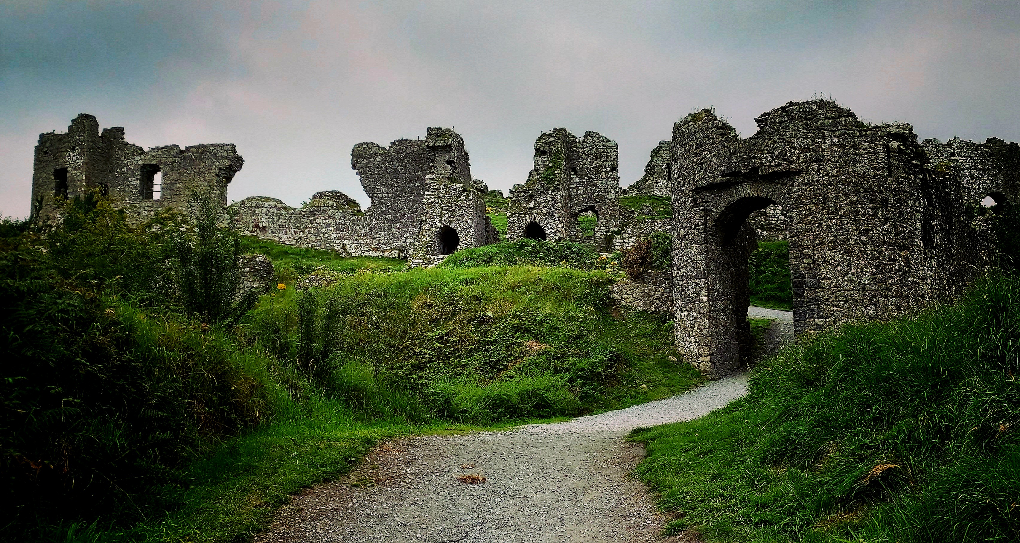 Path among green feields leading to ancient tumbledown castle with various parts leaning at crazy angles silhouetted against cloudy sky.