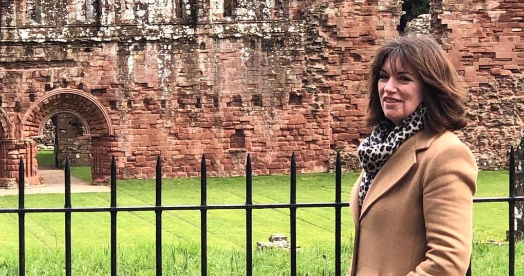A woman with shoulder length brown hair wearing a camel clouored coat stands smiling in front of a black iron railing. Behind here is a larged, ruined sandstone building, the rmains of an abbey, with a decrotative arched adoorway to the left of the picture.